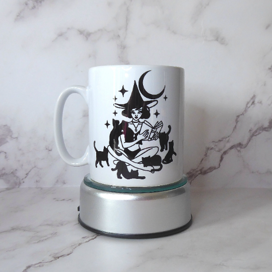 Shows a glossy ceramic mug with a black image of a witch surrounded by black cats