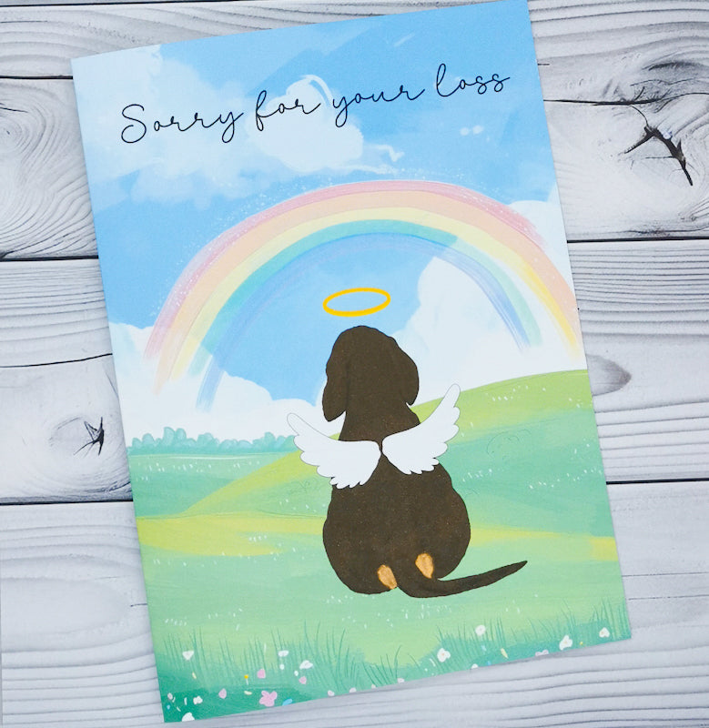 Shows a greeting card which says sorry for your loss and has a dog with wings and a halo sat in a lush green field with a rainbow on the horizon 