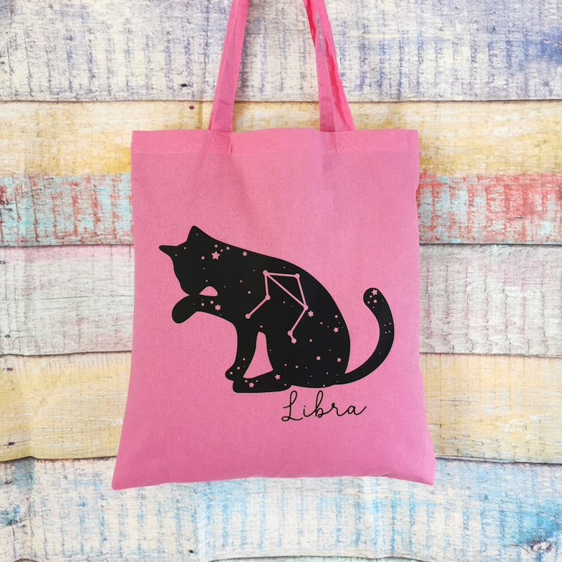 photo shows a pink tote bag which depicts a cat washings its paw in the shape of the libra zodiac, the bag would make a great gift for any cat lover or stargazing enthusiast