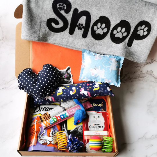 Picture shows a large cat gift box with a personalised grey blanket with the name "Snoop", cat treats, a catnip kicker pillow and heart and various cat toys