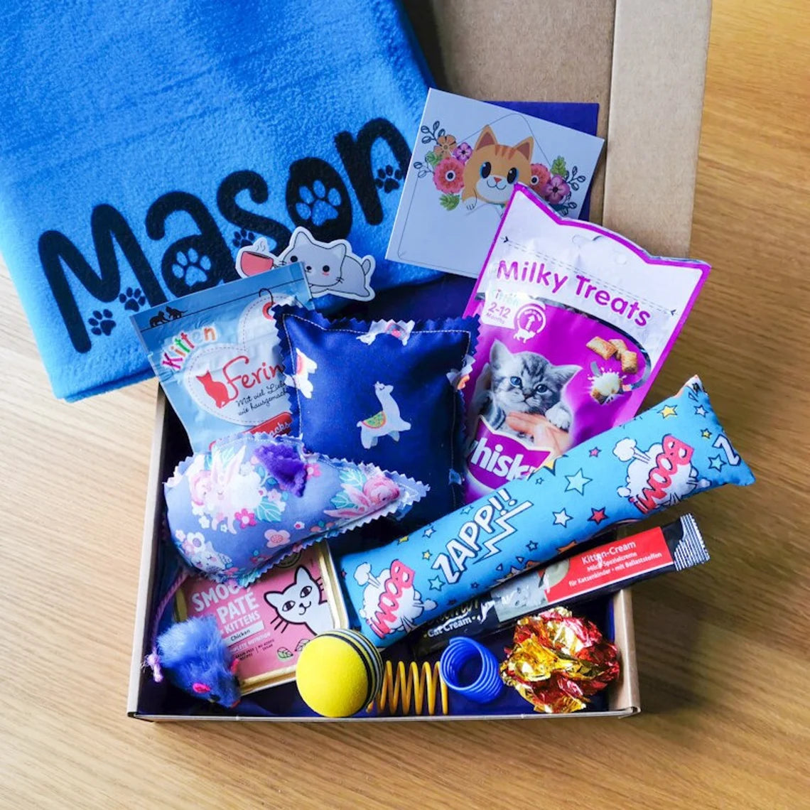 Picture shows a kitten gift box with a personalised blue blanket with the name "Mason", two pouches of cat treats, a cat food tin, a catnip kicker pillow and heart and various cat toys