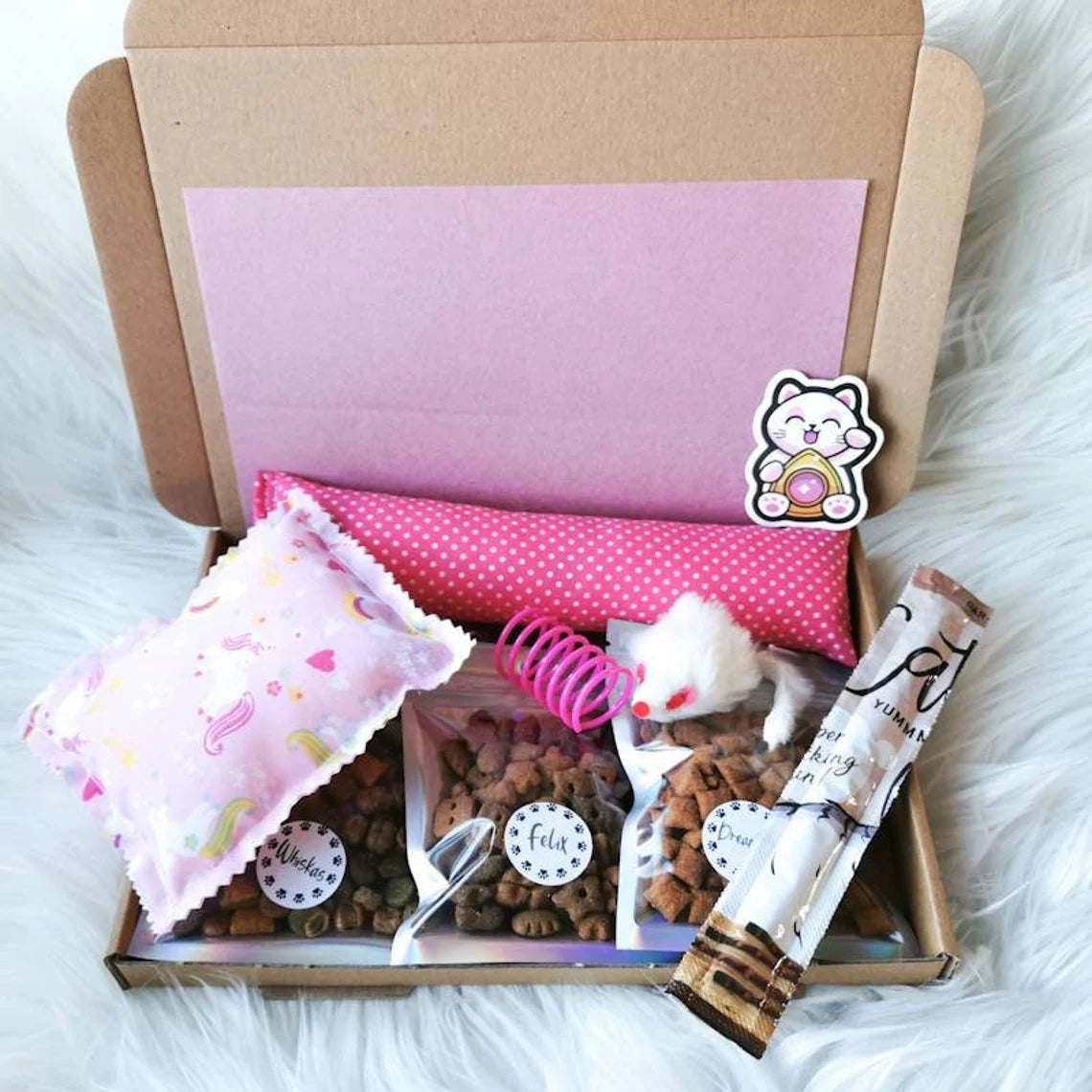 Picture shows a small cat treat gift box in a pink theme, with three small packs of cat treats, a catnip kicker and pillow, a cat treat stick and two toys