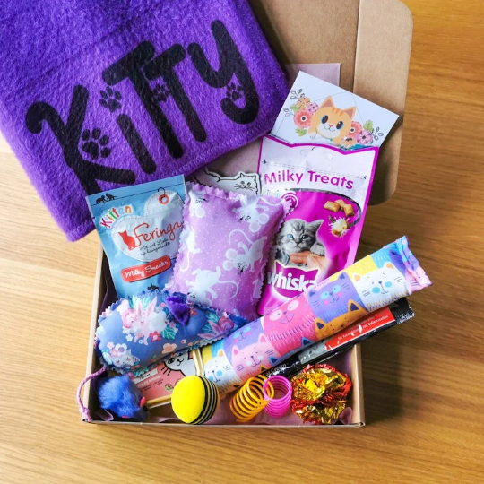 Picture shows a kitten gift box with a personalised purple blanket with the name "Kitty", two pouches of cat treats, a cat food tin, a catnip kicker pillow and heart and various cat toys