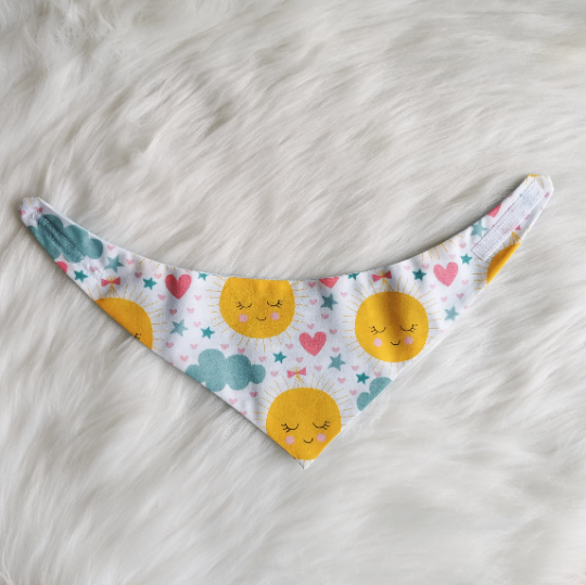 Picture shows a small summer print bandana with smiling sun faces and hearts and clouds, the bandana is in the round the neck style