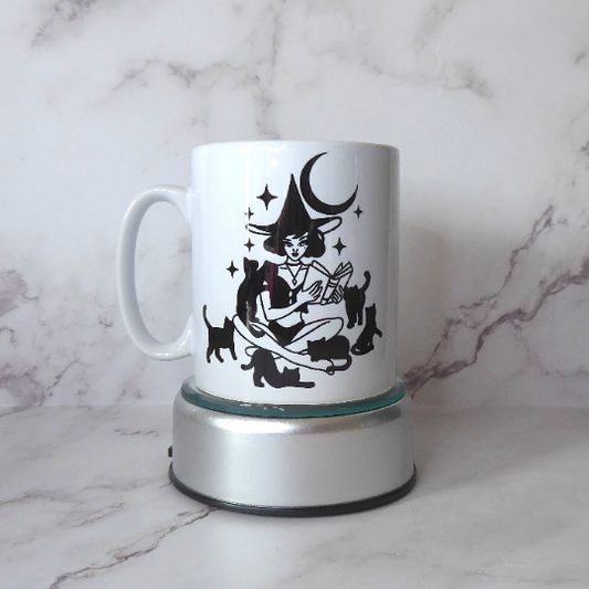 Shows a glossy ceramic mug with a black image of a witch surrounded by black cats 