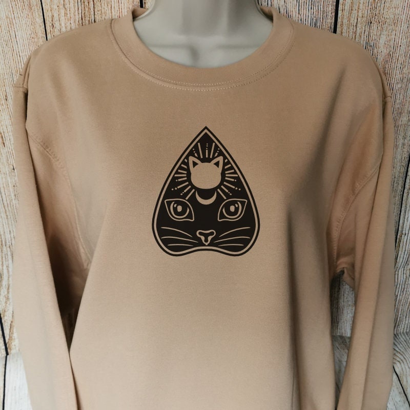 a sweatshirt for cat owners and horror lovers in the colour nude which has a cats head in the shape of a planchette used for ouija boards