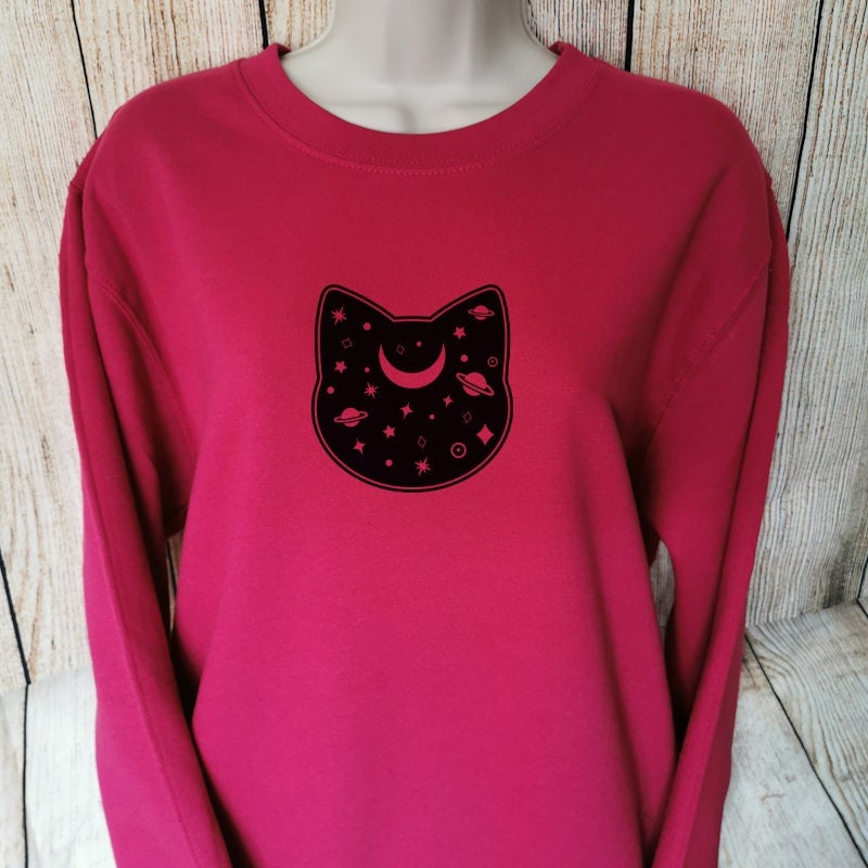 A sweatshirt in the colour cranberry which has a cats head on it, the head is filled with planets, the sweatshirt is great for Cat Owners