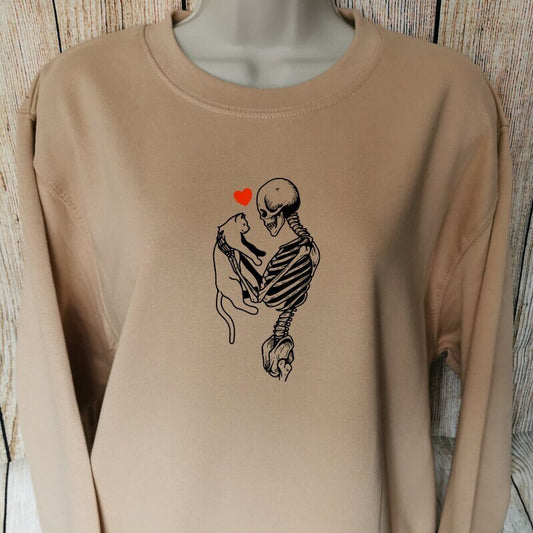 a sweatshirt for cat and horror lovers in the colour nude which shows a skeleton holding a cat with a red heart above them