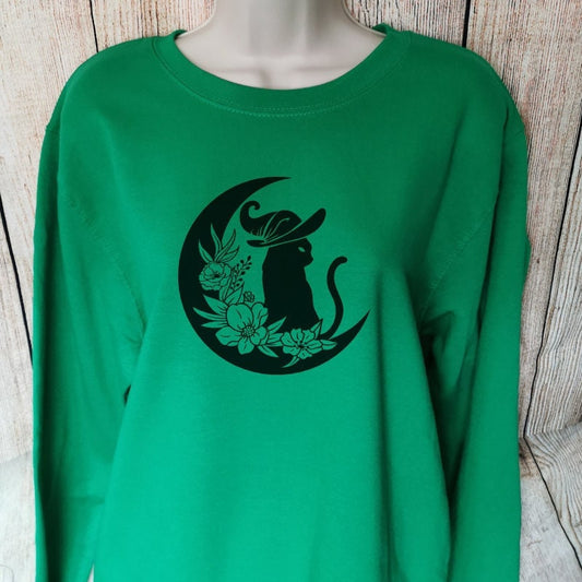 A green sweatshirt that has a black cat in a witches hat, sat in a moon with flowers on, great gift for both cat lovers and witches alike