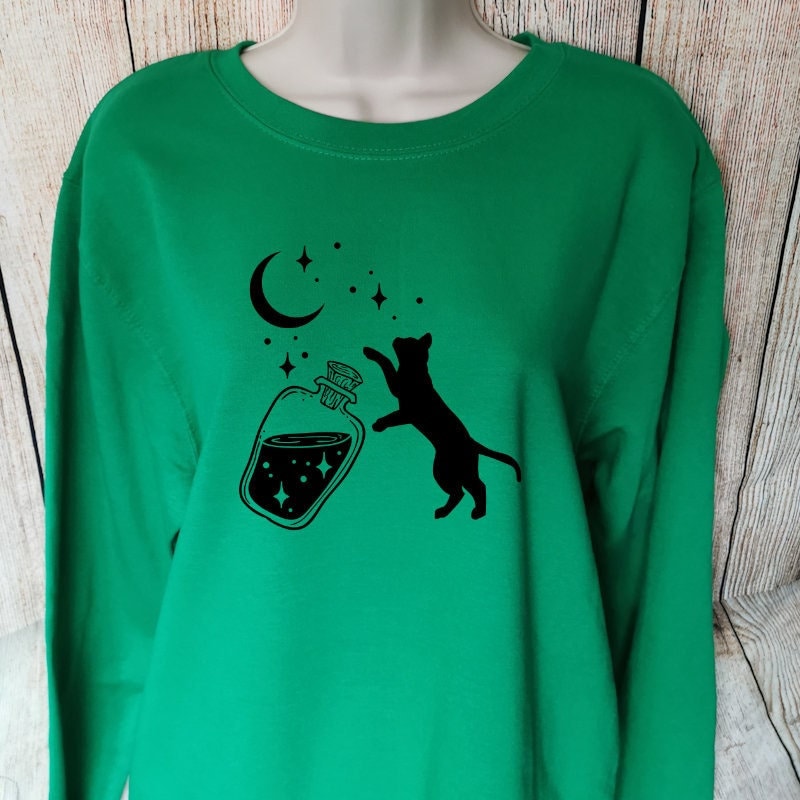 a green sweatshirt which has a black cat chasing stars that have come out of a bottle, the whimsical design makes this a perfect gift for cat lovers and fantasy lovers alike