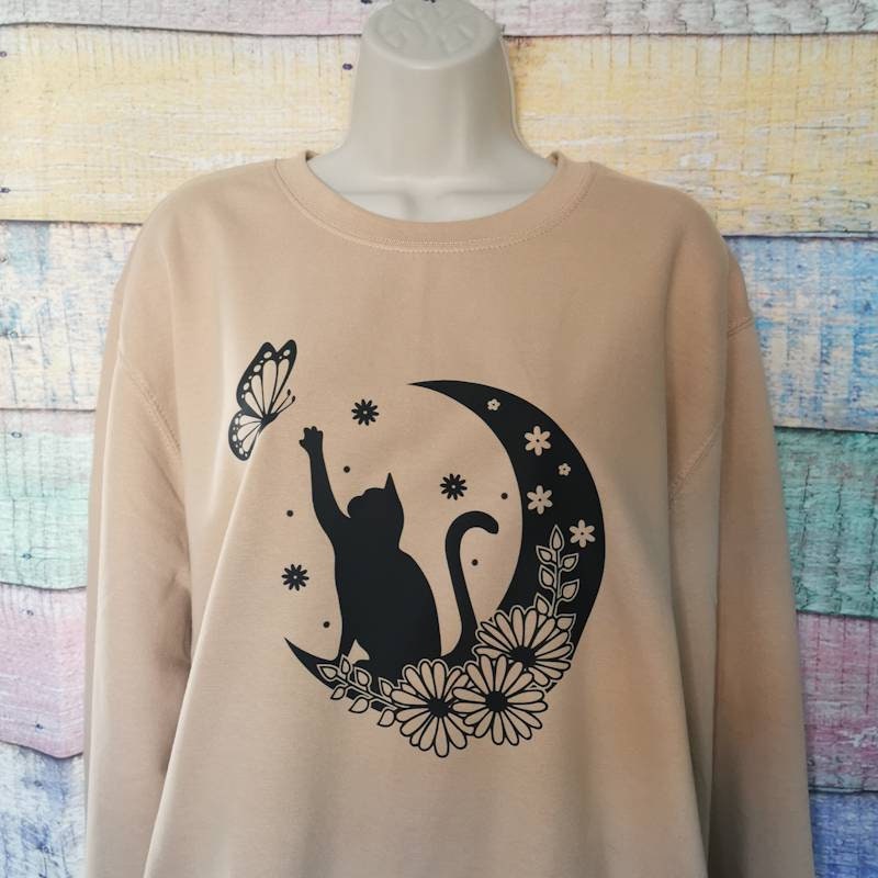 a sweatshirt in the colour nude which has a cat on it reaching for a butterfly while sat on a floral moon, this adorable design is perfect for cat lovers