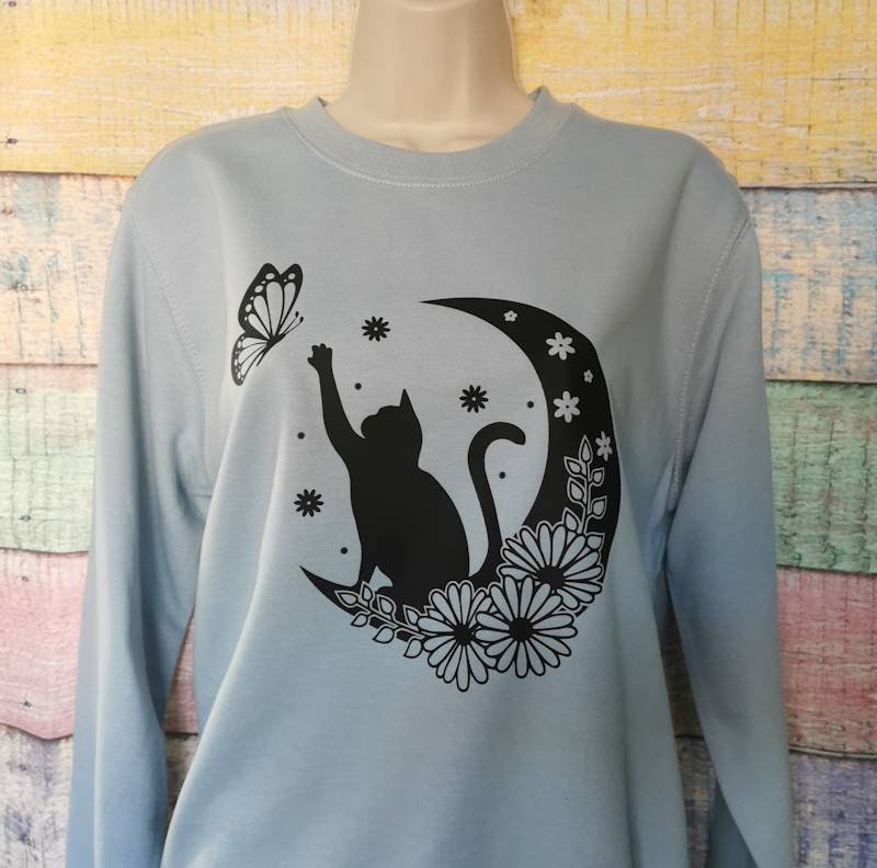 a sweatshirt in the colour light blue which has a cat on it reaching for a butterfly while sat on a floral moon, this adorable design is perfect for cat lovers